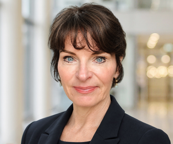 A woman with short brown hair, blue eyes, and a black blazer stands indoors with a blurred office background.