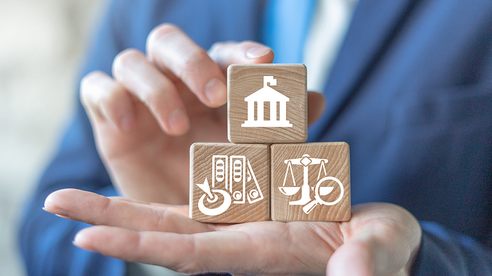 A person in a business suit holds three wooden blocks with icons representing a bank, contracts, and law balance in their hand.