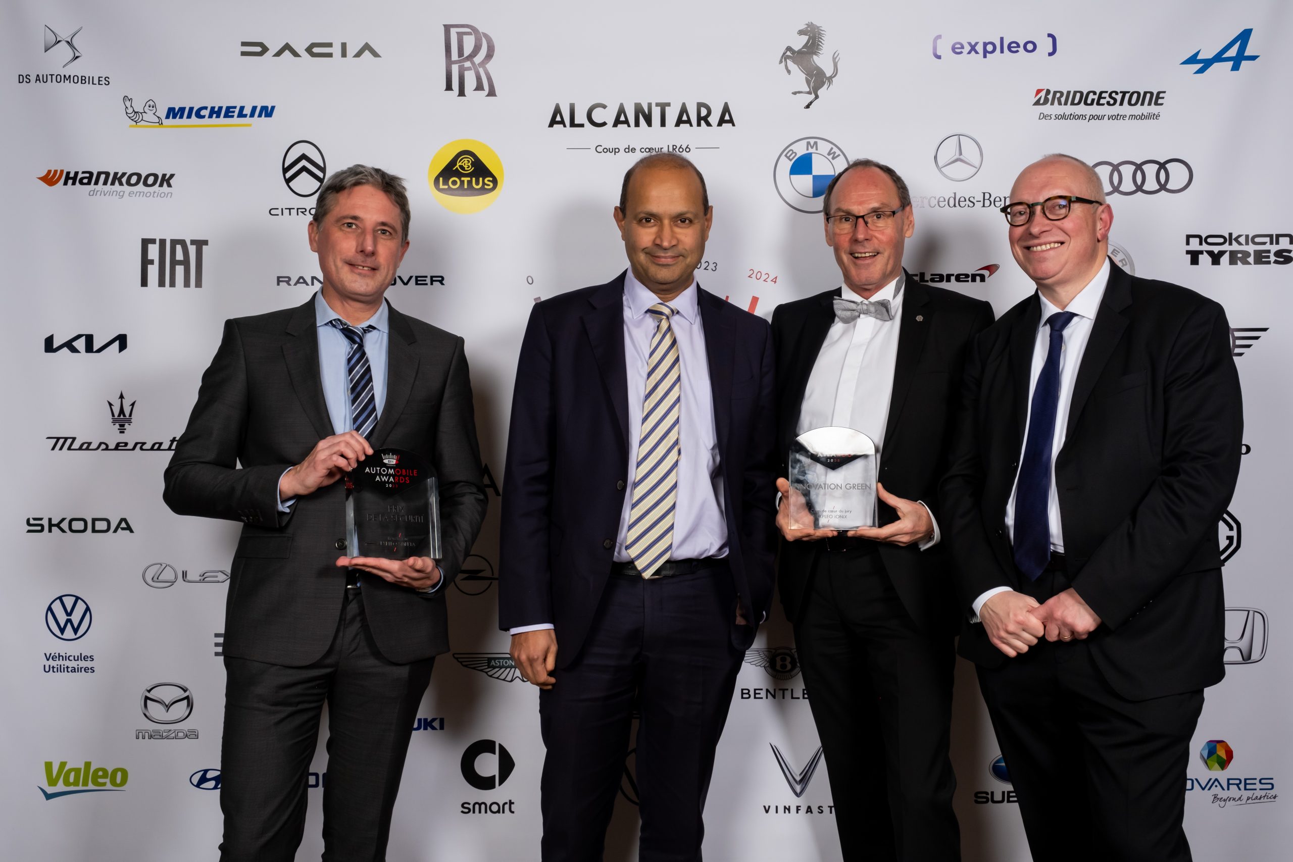 Four businessmen holding awards pose together, smiling at the Expleo Innovations Automobile Awards event, with logos of automotive companies and sponsors in the background.