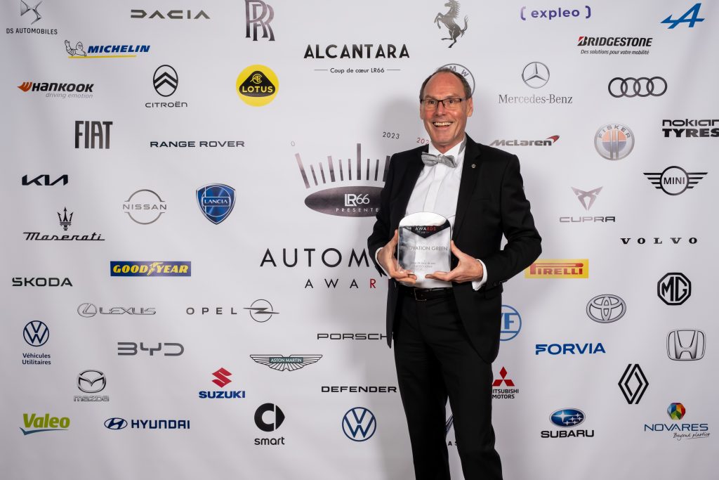 A man in a black tuxedo and bow tie smiling and holding a trophy, standing in front of a backdrop with various automotive brand logos at the Expleo Automobile Awards.