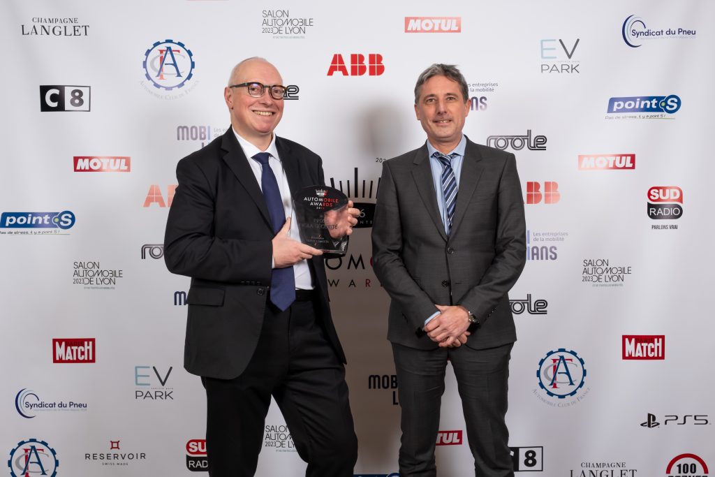 Two men in business suits smiling and posing with an award at the Automobile Innovations Awards, standing in front of a backdrop with multiple corporate logos.