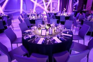 A banquet hall with round tables covered in dark tablecloths, set with plates, glasses, menus, napkins, and floral centerpieces, illuminated by purple lighting. Empty chairs are arranged around each table.