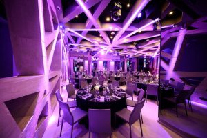 A modern event space with geometric ceiling beams and purple lighting. Round tables are set for dinner with black tablecloths, glassware, and silverware. The room has a futuristic ambiance and mirrored walls.
