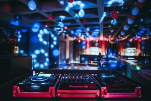 A DJ booth with Pioneer DJ equipment in a dimly lit club, colorful spotlights and balloons hanging from the ceiling, and blurred figures in the background.