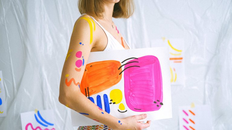 A person in a white tank top holds a colorful abstract painting, reminiscent of the vibrant and creative solutions offered by Expleo services. The painting features vibrant blobs of pink, orange, yellow, and blue. The person has similar colorful paint splatters on their arms. The background showcases sheets with matching abstract art.