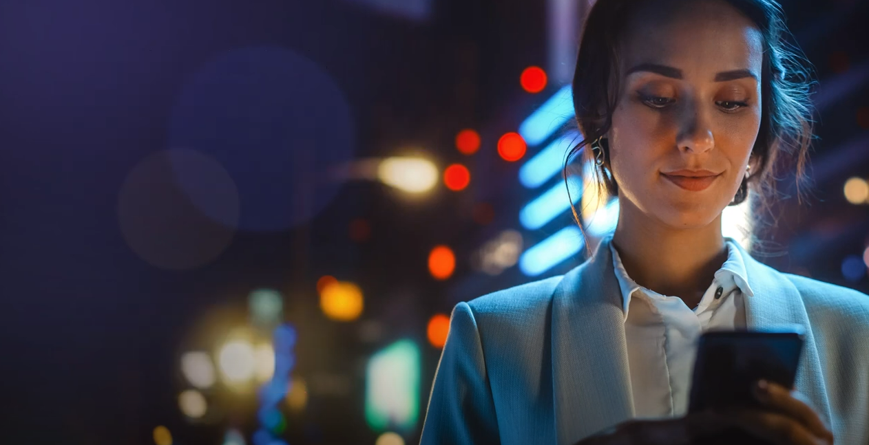 A woman looks at her smartphone at night, illuminated by the screen's light with colorful city lights blurred in the background.