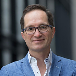 Professional headshot of Peter Hinssen,  wearing glasses, a blue blazer, and a white shirt, smiling at the camera with an urban backdrop.