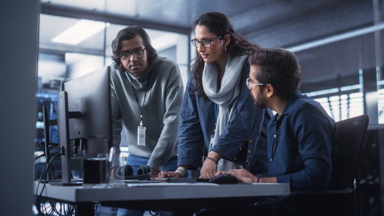 Three people in an office setting are gathered around a desk, looking intently at a computer monitor. Two are standing, one is sitting and typing, and all appear to be collaborating on a project.