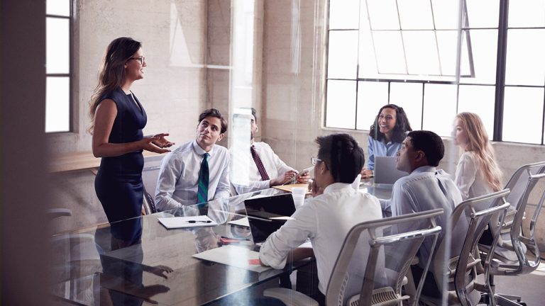 A businesswoman is standing and addressing a group of five colleagues seated around a glass conference table in a modern office with large windows. The attendees are attentively listening, and some have laptops and documents in front of them.