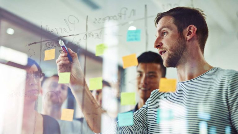 A man writes on a glass board with a marker in an office setting, surrounded by colleagues and sticky notes in the background, suggesting a collaborative work environment.