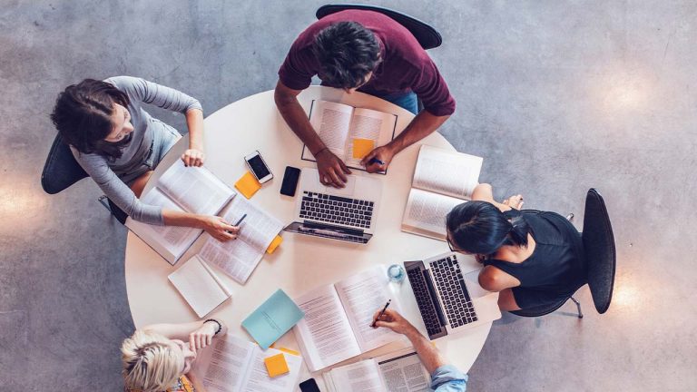 Top view of a diverse group of five students studying together around a round table, with books, laptops, and smartphones visible.