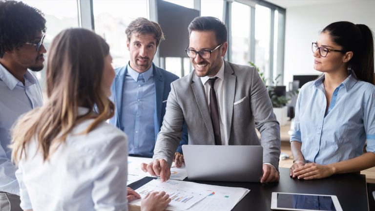 A group of five professionals are engaged in a discussion in a bright office. One man in a gray suit is holding a laptop while another person shows charts and graphs. Large windows in the background let in natural light. They appear focused and collaborative.