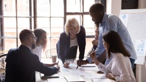 A diverse group of six people are gathered around a table in a modern office. The group is engaged in a collaborative discussion. One person is standing and pointing at documents on the table, while the others are seated, listening and taking notes.