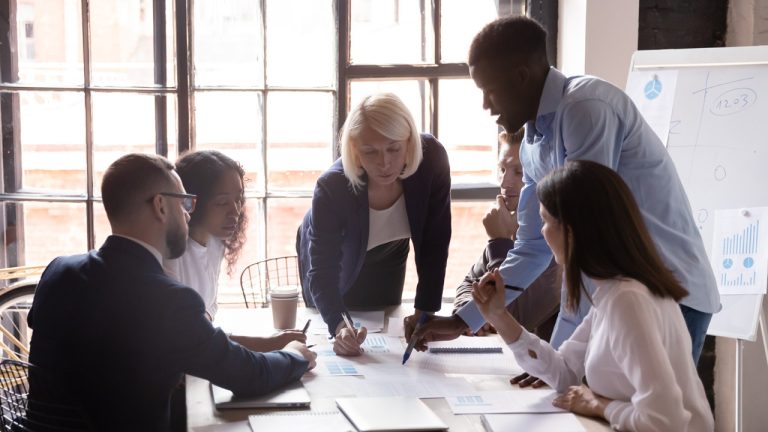 A diverse group of six people are gathered around a table in a modern office. The group is engaged in a collaborative discussion. One person is standing and pointing at documents on the table, while the others are seated, listening and taking notes.