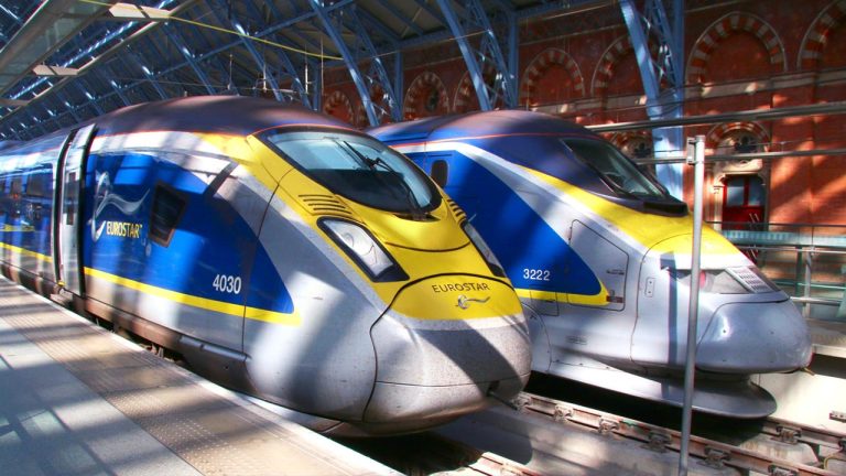 Two Eurostar trains are parked side by side at a station. Both trains, adorned in blue, yellow, and silver, display the Eurostar logo. The background features an arched roof and brick building. Sunlight illuminates the modern, sleek design of the trains.
