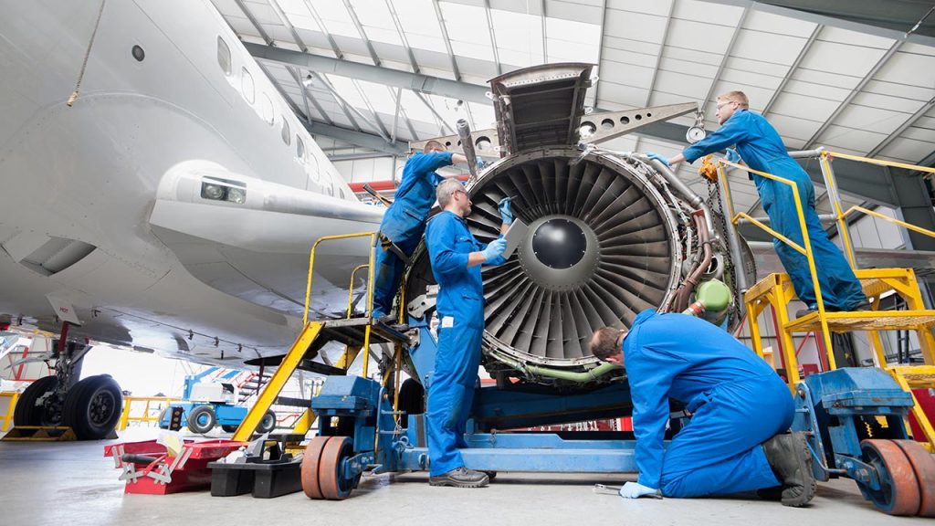 Four engineers in blue coveralls inspect and repair a large jet engine inside an aircraft hangar, using tools and ladders around the exposed engine while the airplane is parked indoors.