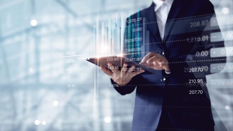 A person in a business suit uses a tablet, with floating holographic financial graphs and data projections emerging from the device, indicating analysis or financial management. The background consists of blurred blue and gray tones, suggesting a modern office environment.