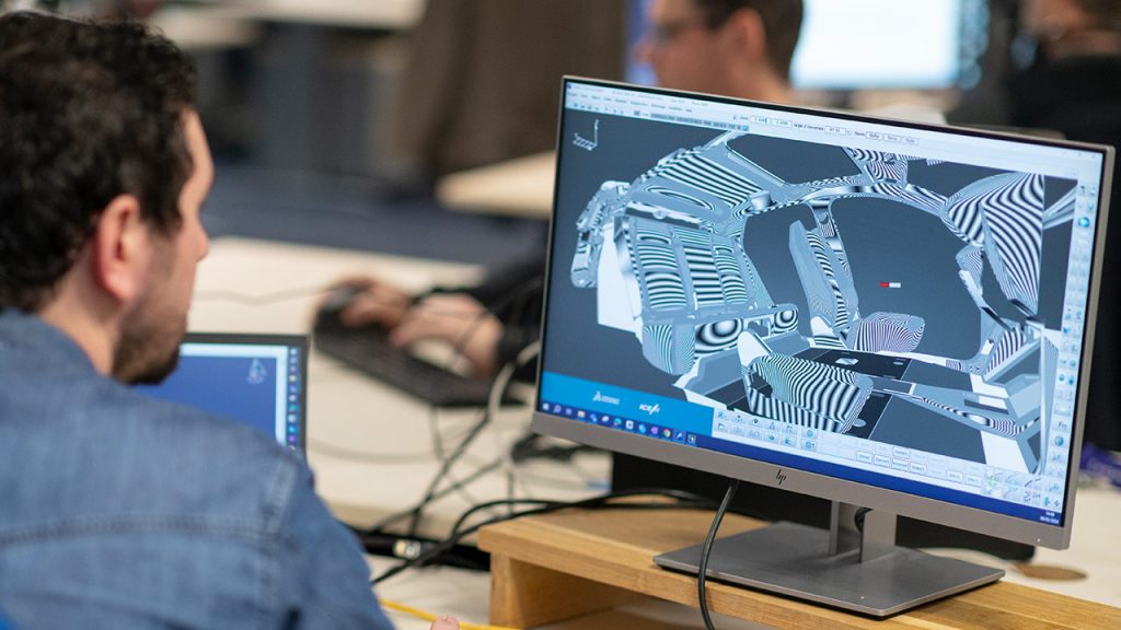 A person sits at a desk working on a 3D design using computer software. The design on the monitor appears to be a complex structure with overlapping patterns. Another person works at a computer in the background. The workspace is equipped with computers and tech gadgets.