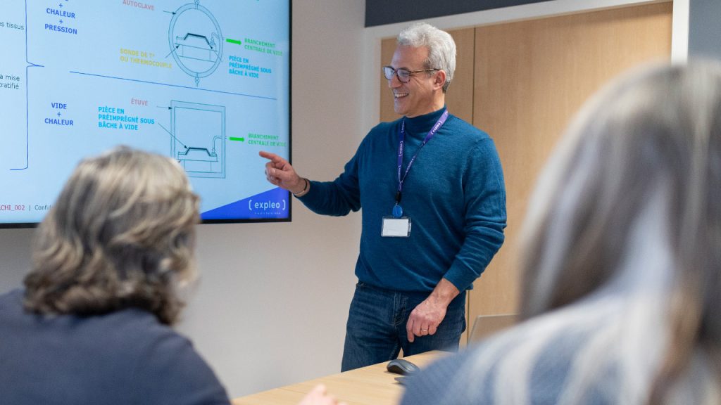 A man with gray hair and glasses is giving a presentation in a conference room, pointing to a diagram on a screen. Two people are seated at the table, observing. The presentation appears to involve technical or engineering concepts.