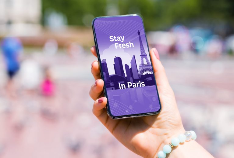 A hand holding a smartphone displays a purple-themed graphic with illustrations of Parisian landmarks, including the Eiffel Tower. The text on the screen reads, "Stay Fresh In Paris." The background is slightly blurred, with people and pigeons visible.