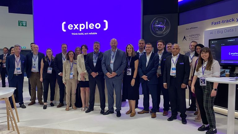 A group of people stands together for a photo in front of a large display screen with the text "{expleo} Think bold, act reliable." Everyone in the group is wearing business attire and name badges. The setting appears to be a professional event or conference.