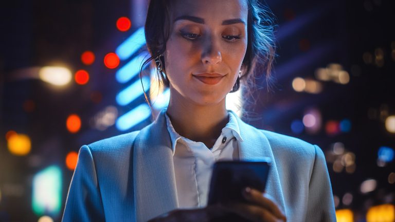 A woman in a white blouse and light blazer stands outdoors at night, looking down and smiling at her phone. The background is illuminated with colorful city lights and vibrant neon signs, creating a lively urban atmosphere.
