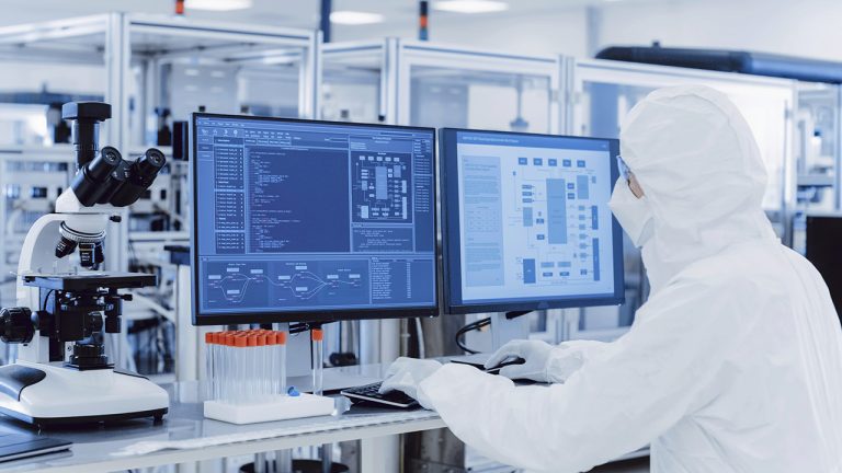 A scientist in full protective gear works at a computer station in a laboratory. The lab setup includes a microscope on the left and multiple test tubes. Two computer monitors display complex scientific data and graphics. Various lab equipment is visible in the background.