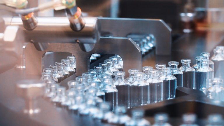 A close-up of a pharmaceutical production line where small glass vials are being automated through a filling machine. The machinery is precisely aligned to fill each of the vials, showcasing a sterile and efficient manufacturing process.