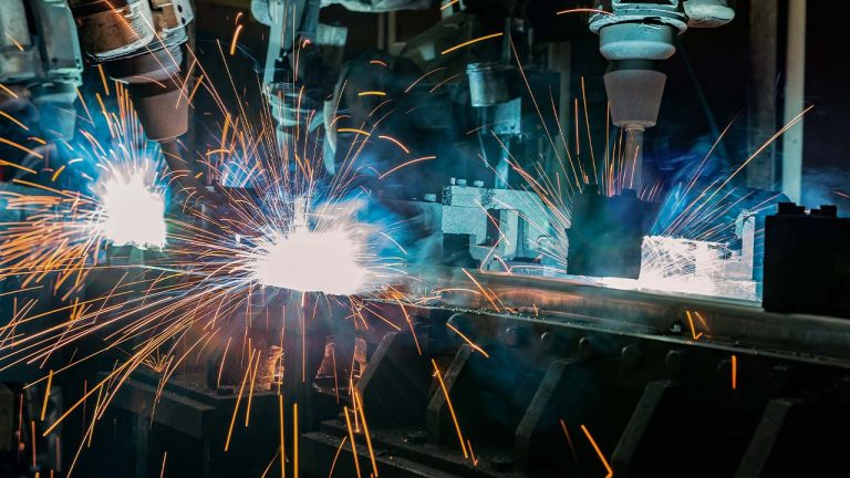Robotic arms in an industrial setting perform welding, emitting bright sparks and smoke. The scene is focused on automation and manufacturing processes, highlighting precision and technology in action.