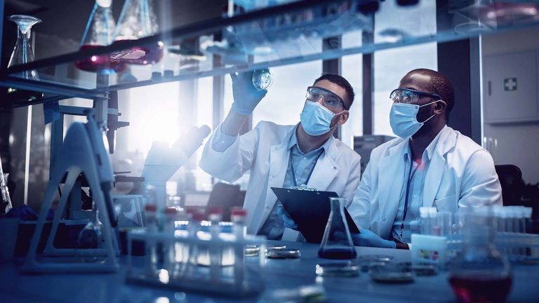 Two scientists in a laboratory analyze a petri dish, wearing masks, gloves, and lab coats. The lab is filled with scientific equipment, glassware, and test tubes, with sunlight streaming through a window.