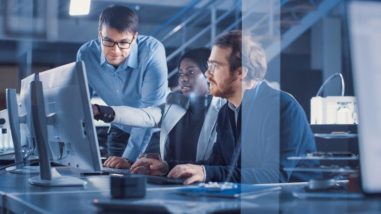 Three people collaborating at a desk with computers. One person is pointing at the screen, while the others observe. The environment looks like a modern office or tech workspace. Glass partitions and staircases are visible in the background.