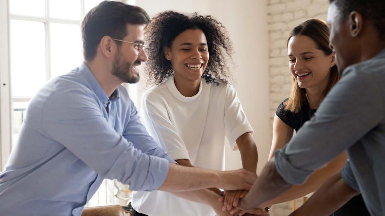 Four colleagues stand in a circle, smiling and stacking their hands together in a gesture of teamwork and unity, in a bright office setting.