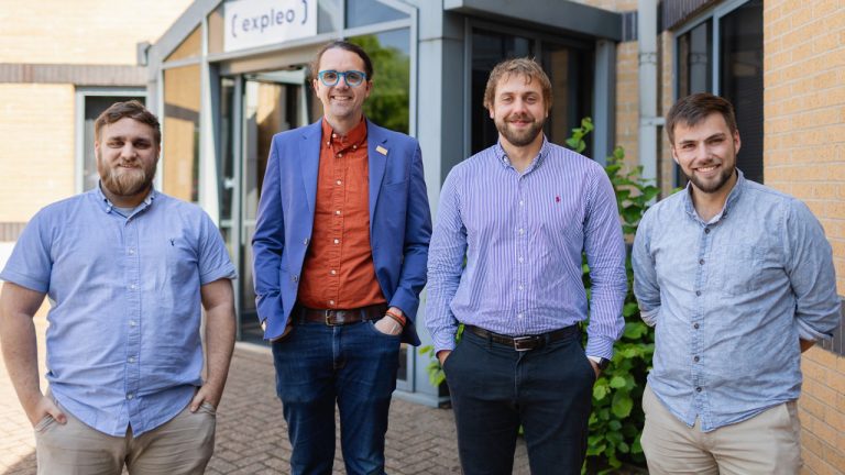 Four men stand and smile outside a building with a sign that reads "expleo." Three wear light blue shirts, and one wears a blue blazer over an orange shirt. They stand on a brick walkway near a green bush.