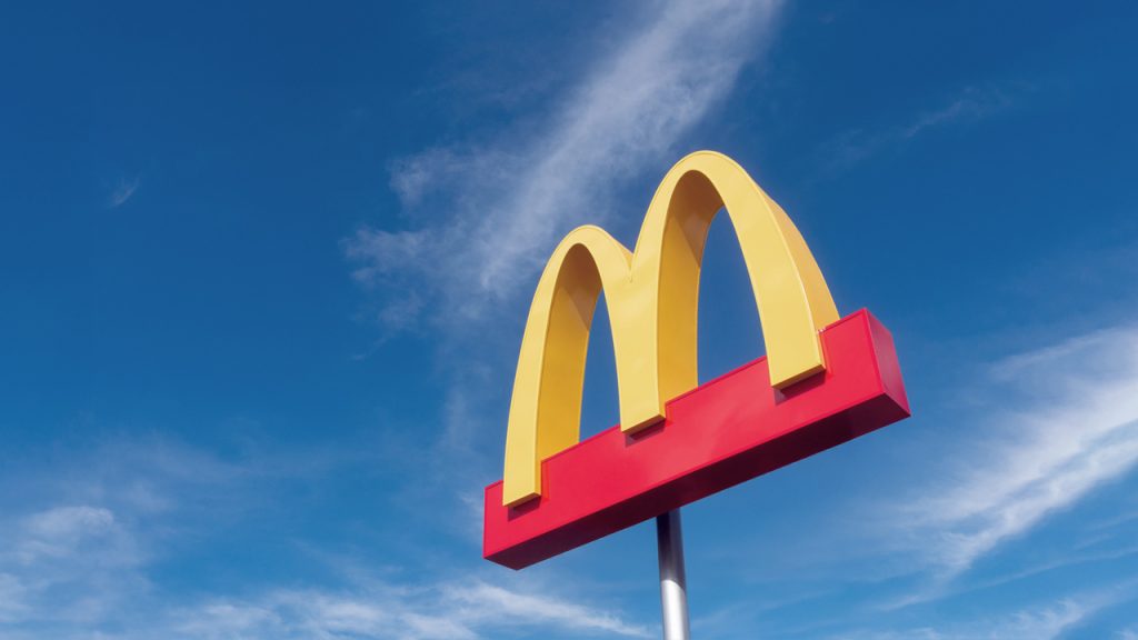 A McDonald’s sign featuring the iconic yellow golden arches logo on a red base, set against a bright blue sky with wispy clouds.