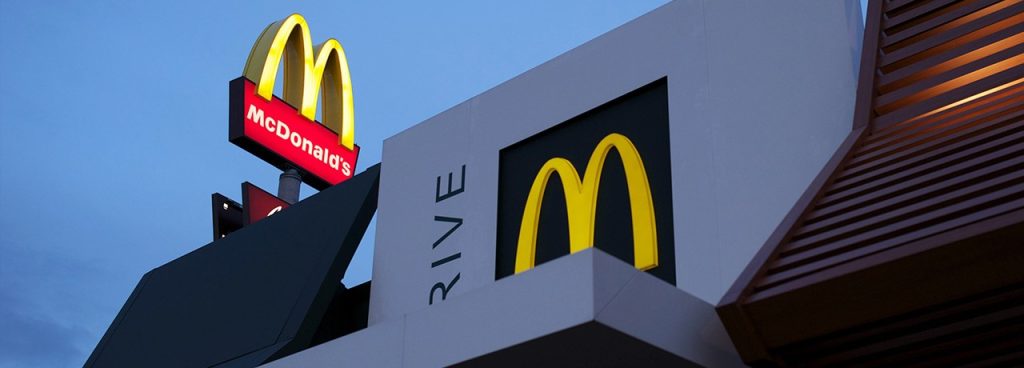 A McDonald's restaurant at dusk, featuring the iconic yellow "M" logo on both a sign and the building, with the word "DRIVE" partially visible. The sky is clear and blue.