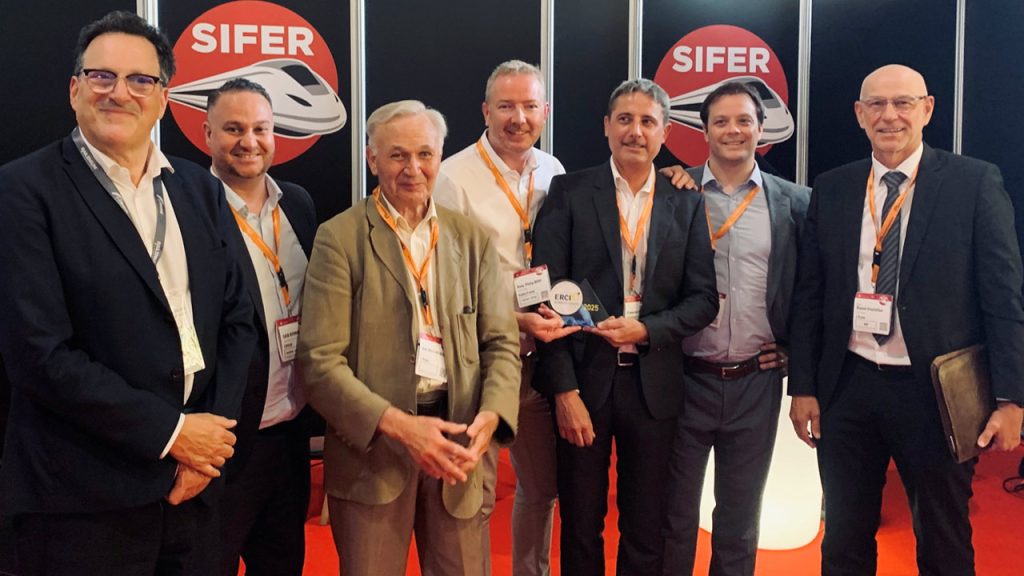 Seven men in business attire stand together, smiling at a SIFER event. One man at the center holds an award. SIFER logos with a train graphic are visible on the wall behind them. All wear orange lanyards.