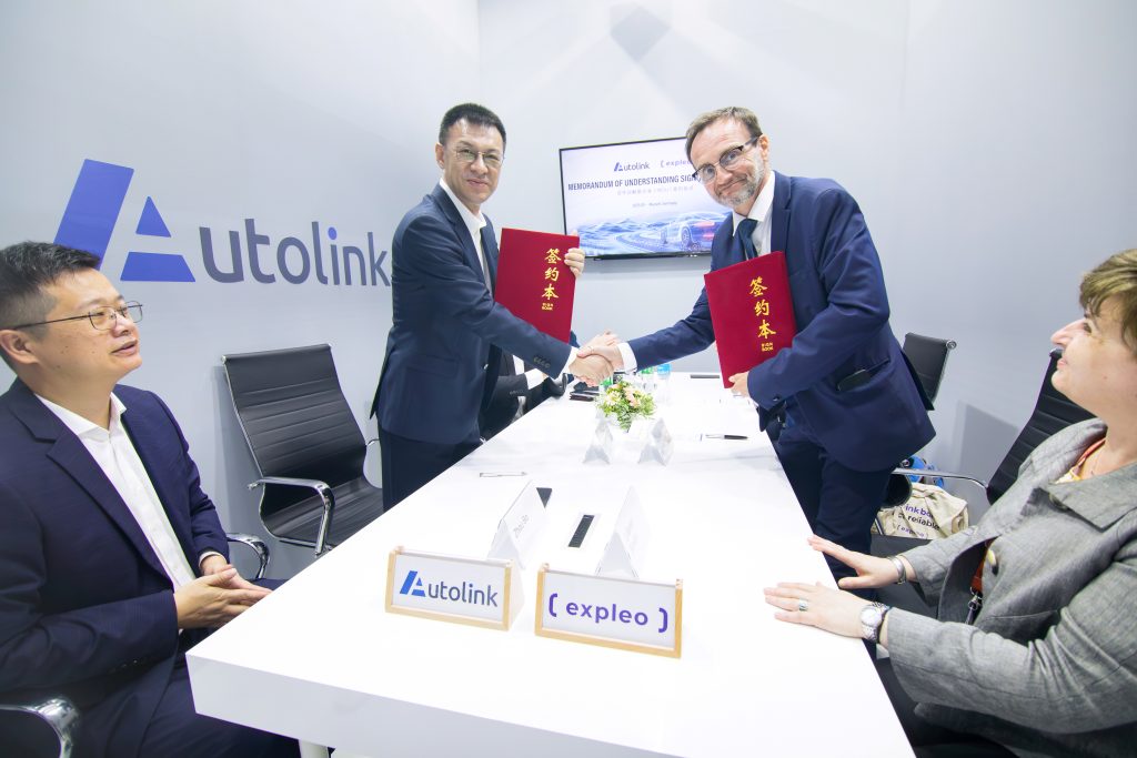 Four business professionals, two standing and shaking hands while holding red folders, and two seated, are gathered around a table with "Autolink" and "Expleo" nameplates, suggesting a formal partnership agreement.
