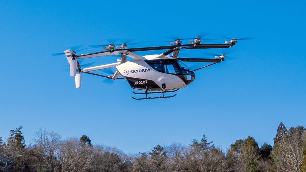 A white SkyDrive flying car, with multiple rotors, hovers in the air above trees against a clear blue sky.