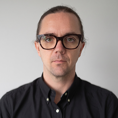 A person with glasses, long hair tied back, and a black button-up shirt, standing against a plain light background, looking directly at the camera with a neutral expression.