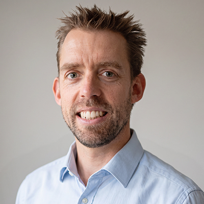 A man with short brown hair and a beard smiles whilst wearing a light blue collared shirt, standing in front of a neutral grey background.