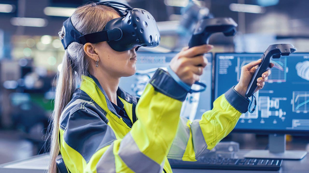 A woman in a high-visibility jacket uses a virtual reality headset and controllers in a modern office with large monitors displaying technical graphics.