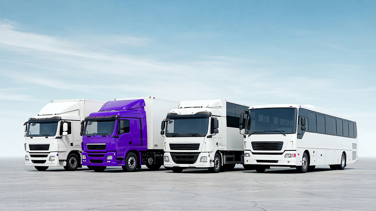 Four large vehicles, including two white trucks, one purple truck, and one white bus, are parked side by side on an open paved area under a clear blue sky.