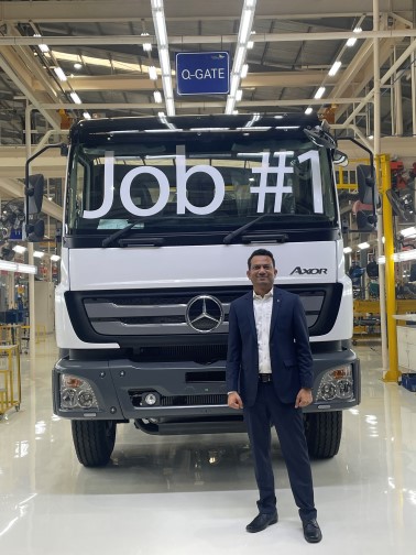 A man in a suit stands smiling in front of a large Mercedes-Benz Axor truck with "Job #1" written on its windshield inside a brightly lit factory.