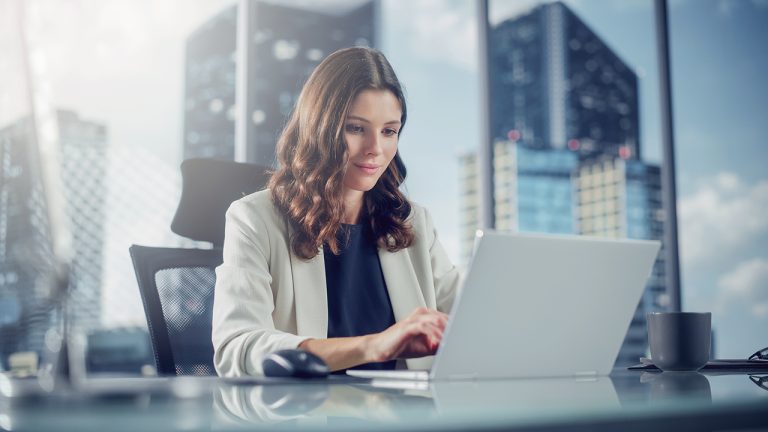 A woman with wavy brown hair wearing a white blazer and dark top works on a laptop at a desk in a modern office with large windows overlooking city skyscrapers.