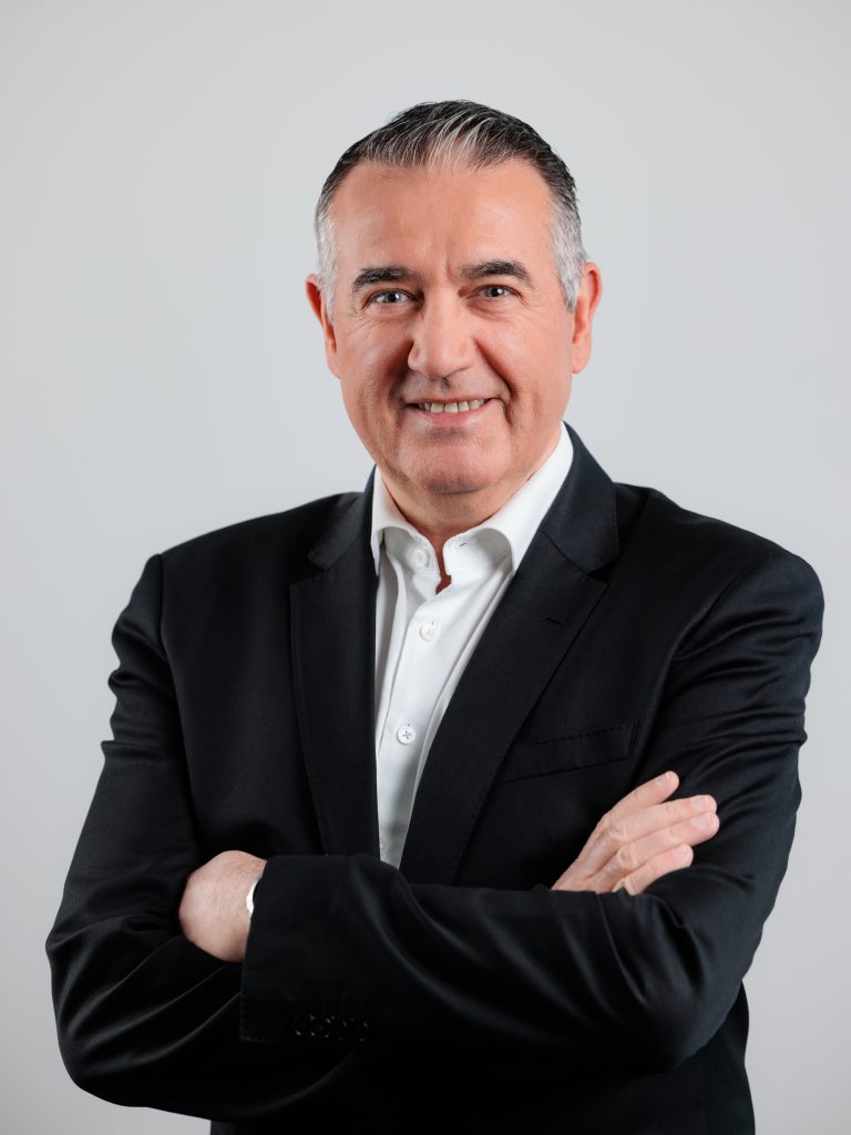 A middle-aged man with gray hair, wearing a black suit and white dress shirt, stands with arms crossed and smiles confidently against a plain light gray background.