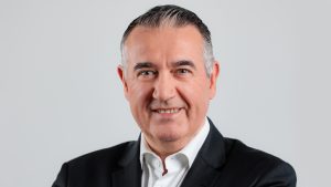 A middle-aged man with short, dark gray hair wearing a black suit jacket and white dress shirt, smiling warmly at the camera against a plain light gray background.