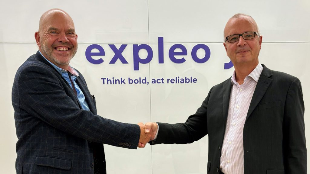 Two men in business attire shake hands and smile in front of a wall with the Expleo logo and the slogan "Think bold, act reliable.