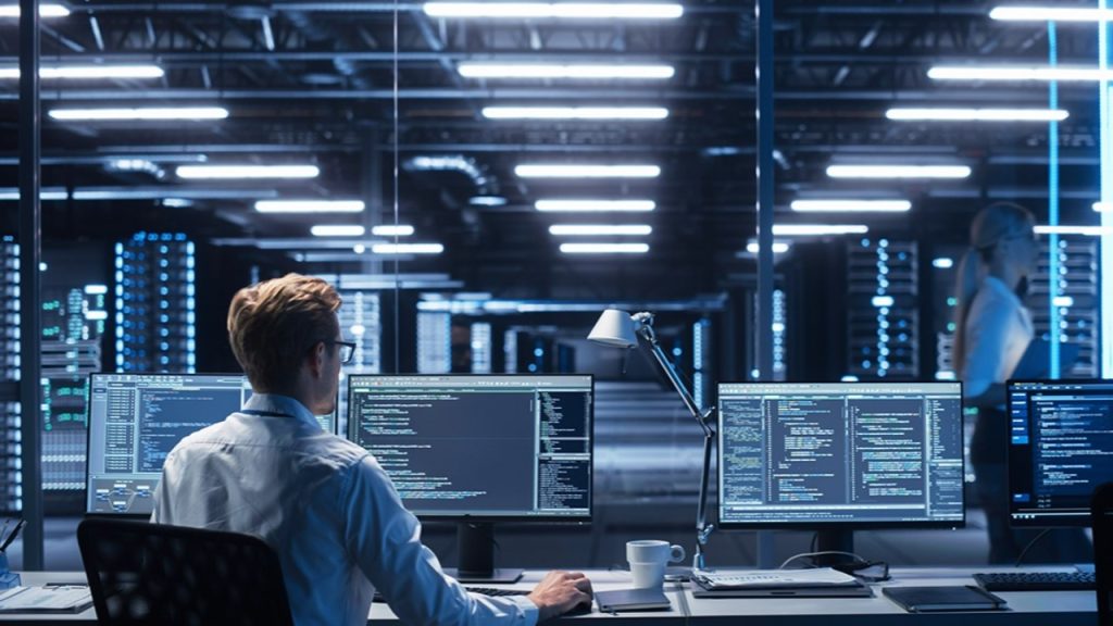 A person sitting at a desk with three monitors displaying code, working in a modern, glass-walled server room filled with rows of servers and bright overhead lighting.