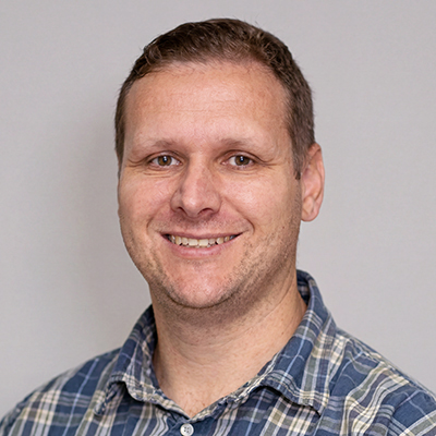 A man with short light brown hair, wearing a blue and white checked shirt, smiles at the camera against a plain light grey background.