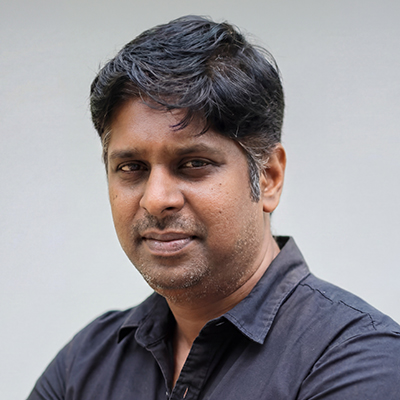 A man with short, dark hair and medium skin tone is wearing a black button-up shirt. He is looking at the camera with a neutral expression against a plain light grey background.